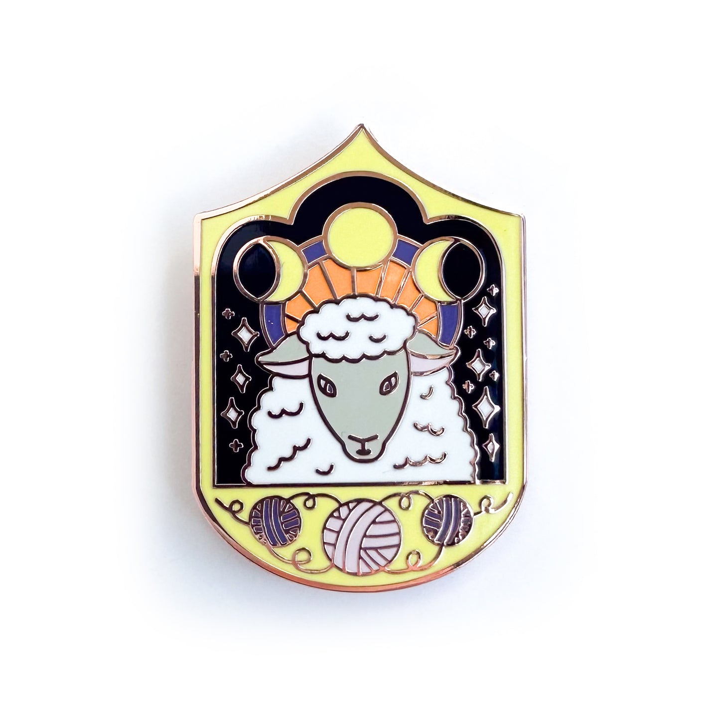An enamel pin with a sheep surrounded by stars in a yellow Fram. The sheep has a halo with moon phases on it and the frame has yarn balls on it. 