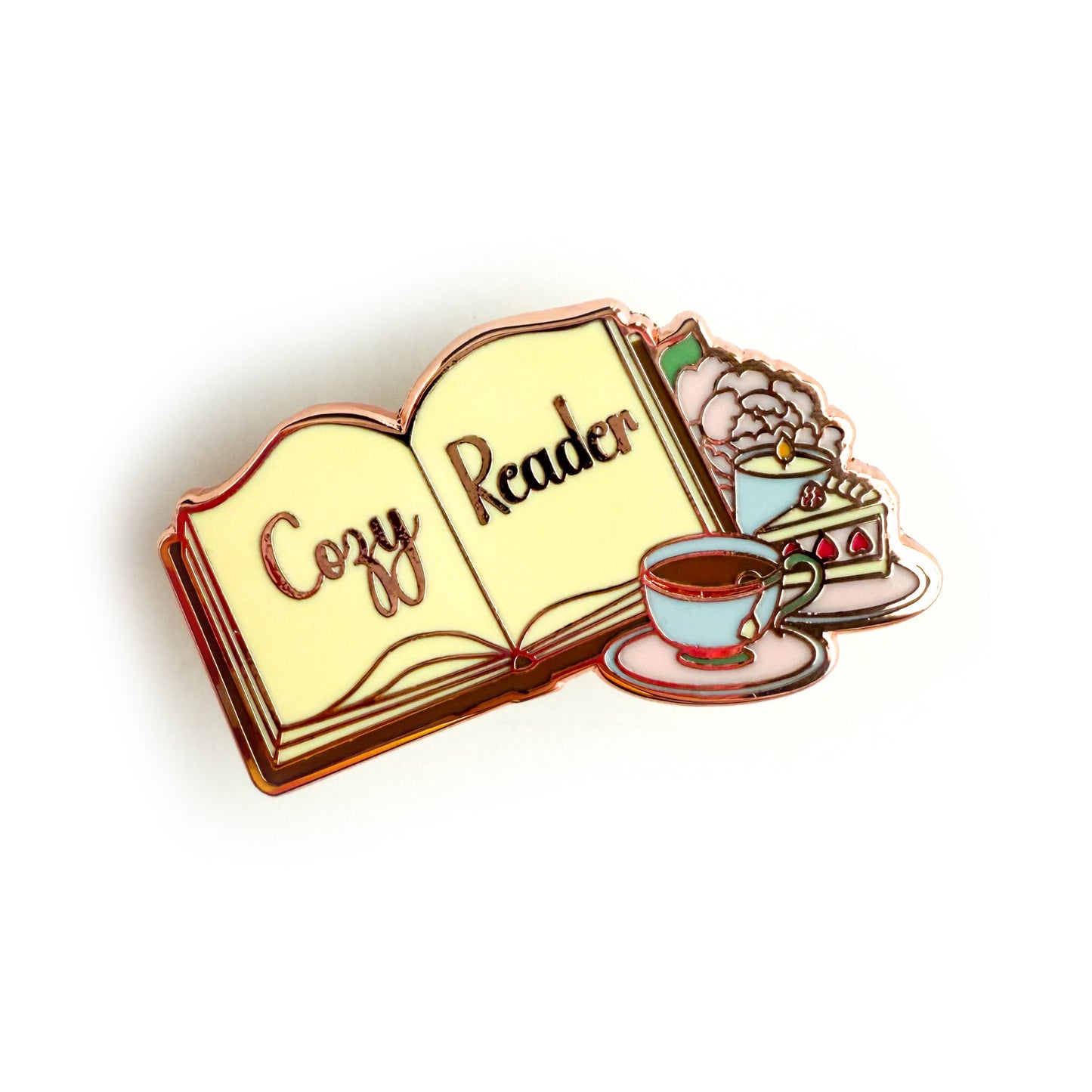 Am enamel pin depicting an open book with “Cozy Reader” written across the pages. Beside the book are a full tea cup on a pink saucer, a slice of strawberry cake, a lit candle, and a baby pink peony with a green leaf.