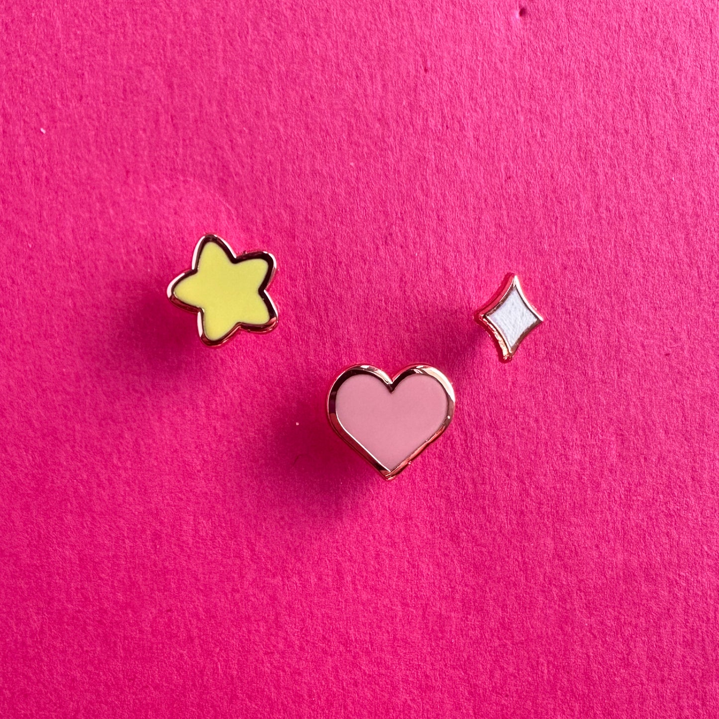 Three mini enamel pins, one is a yellow puffy star, one is a pink heart and one is a diamond sparkle. They are all on a pink paper background.