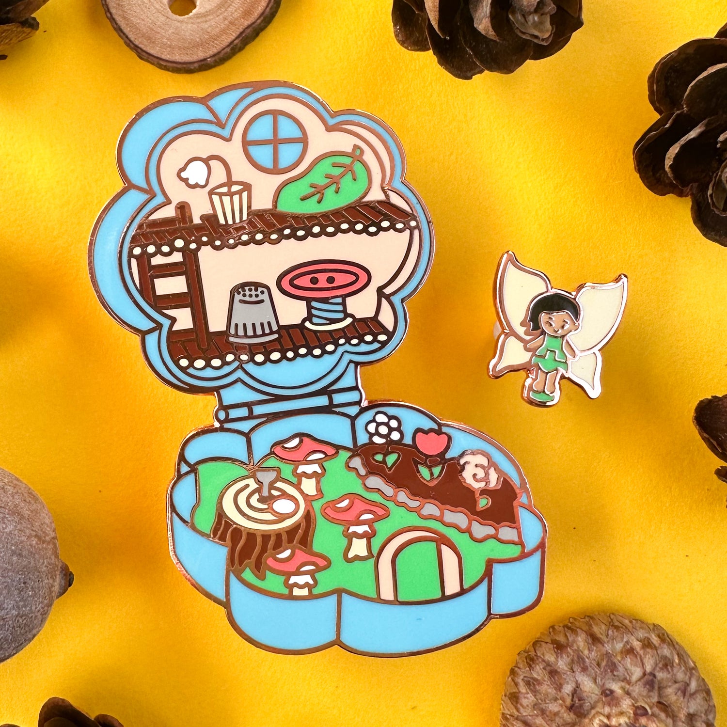 An enamel pin shaped like a Polly Pocket toy with a tiny fairy doll and a flower compact house. The pin is on a yellow background with pinecones and acorns strewn about it. 