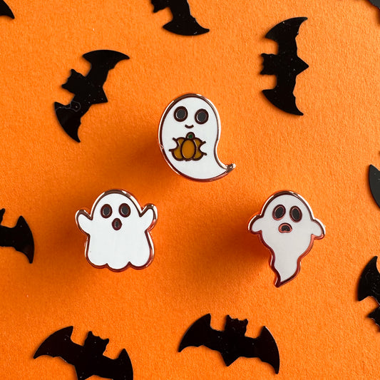 Three small enamel pins each in the shape of a cartoon ghost. They are drawn in a cute art style. The ghost pins are on an orange background with bat shaped confetti around them.