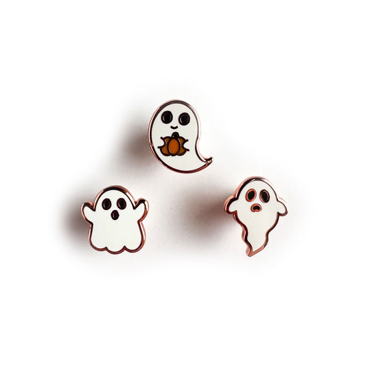 Three small enamel pins each in the shape of a ghost, the middle one is smiling and holding a pumpkin, the left one is making a spooky face, and the right one is frowning.