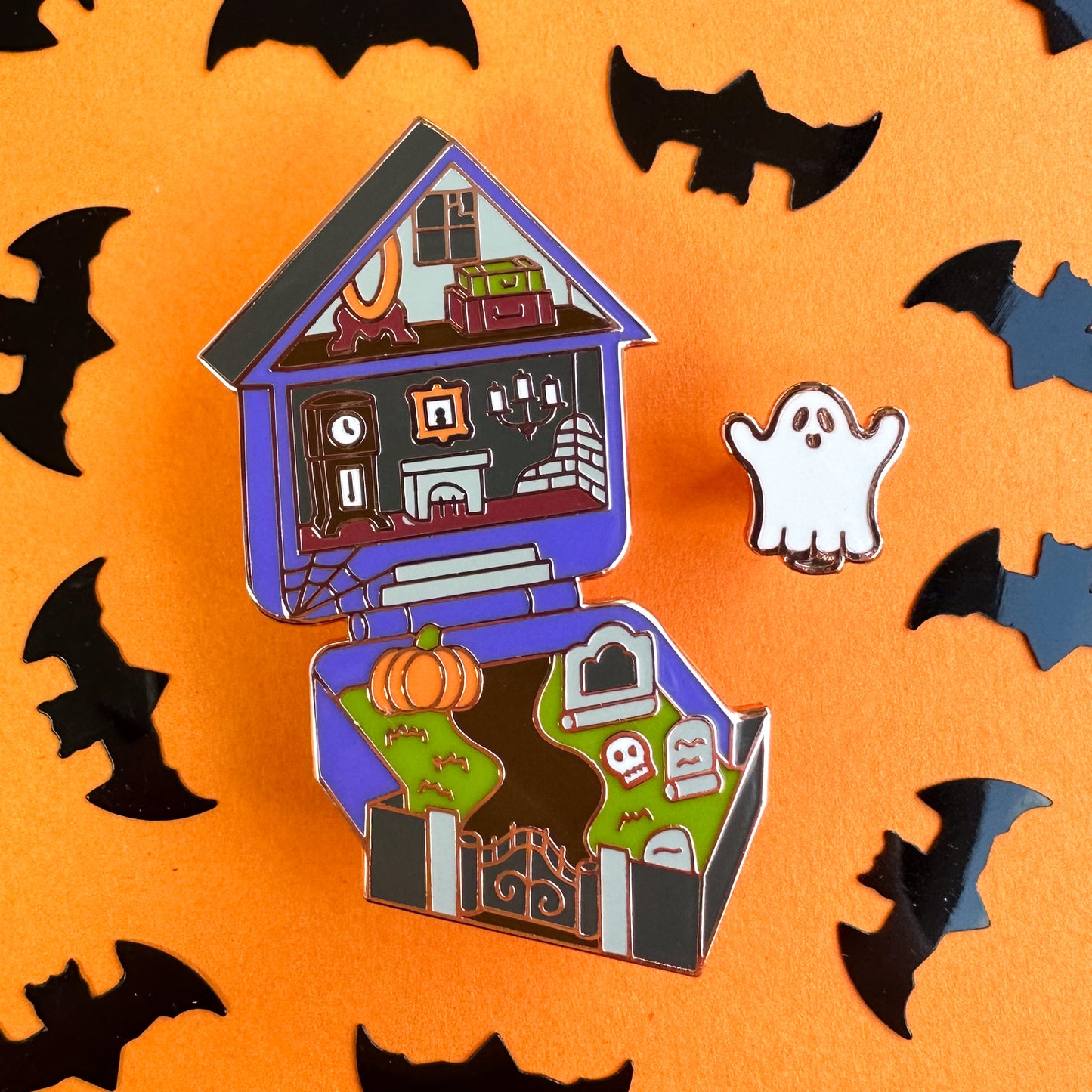 A haunted house and ghost pin set in the style of Polly Pocket toys. The house has a spooky attic with a broken window, a fireplace and a yard featuring a pumpkin and gravestones. The house is purple with a black roof.
