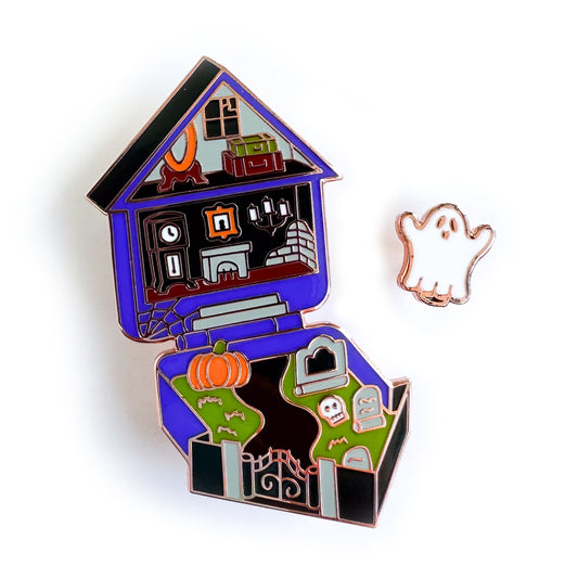 Two enamel pins, one shaped like a little sheet ghost, the other shaped like a Polly Pocket toy house but the house is haunted. It has little details like a broken window, candelabra, and gravestones in the yard.