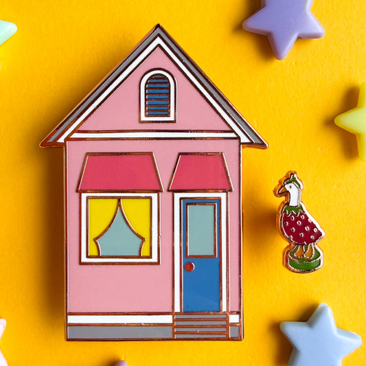 A pink house shaped enamel pin with a blue door and yellow curtains and a mini pin shaped like a porch goose in a berry costume. The pins are hard enamel with copper metal and are on a yellow background with plastics stars around it.