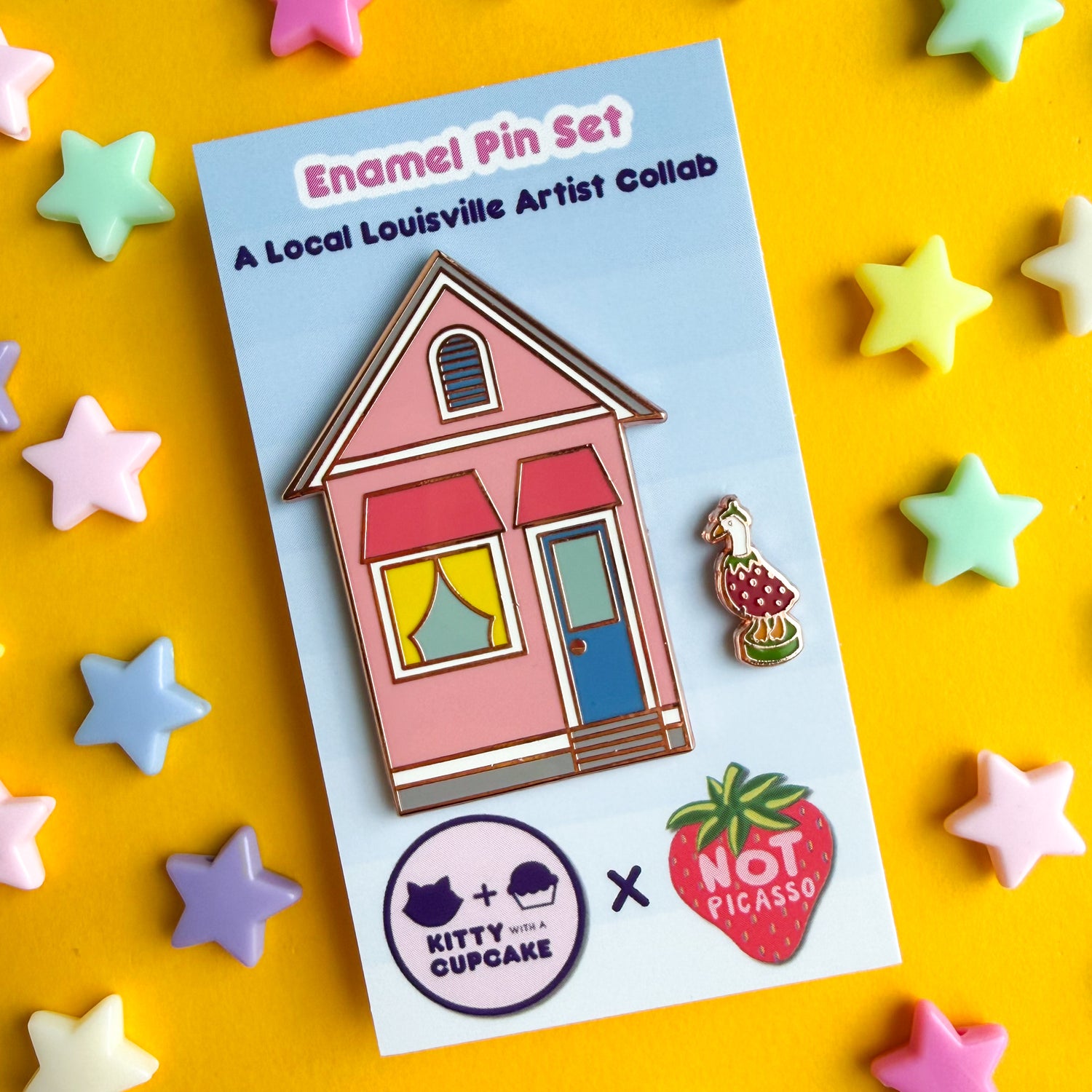A pink house shaped enamel pin with a blue door and yellow curtains and a mini pin shaped like a porch goose in a berry costume. The pins are packaged on a blue card that reads "Enamel Pin Set - A Local Louisville Artist Collab" below the pins are the logos of Kitty With a Cupcake and Not Picasso.