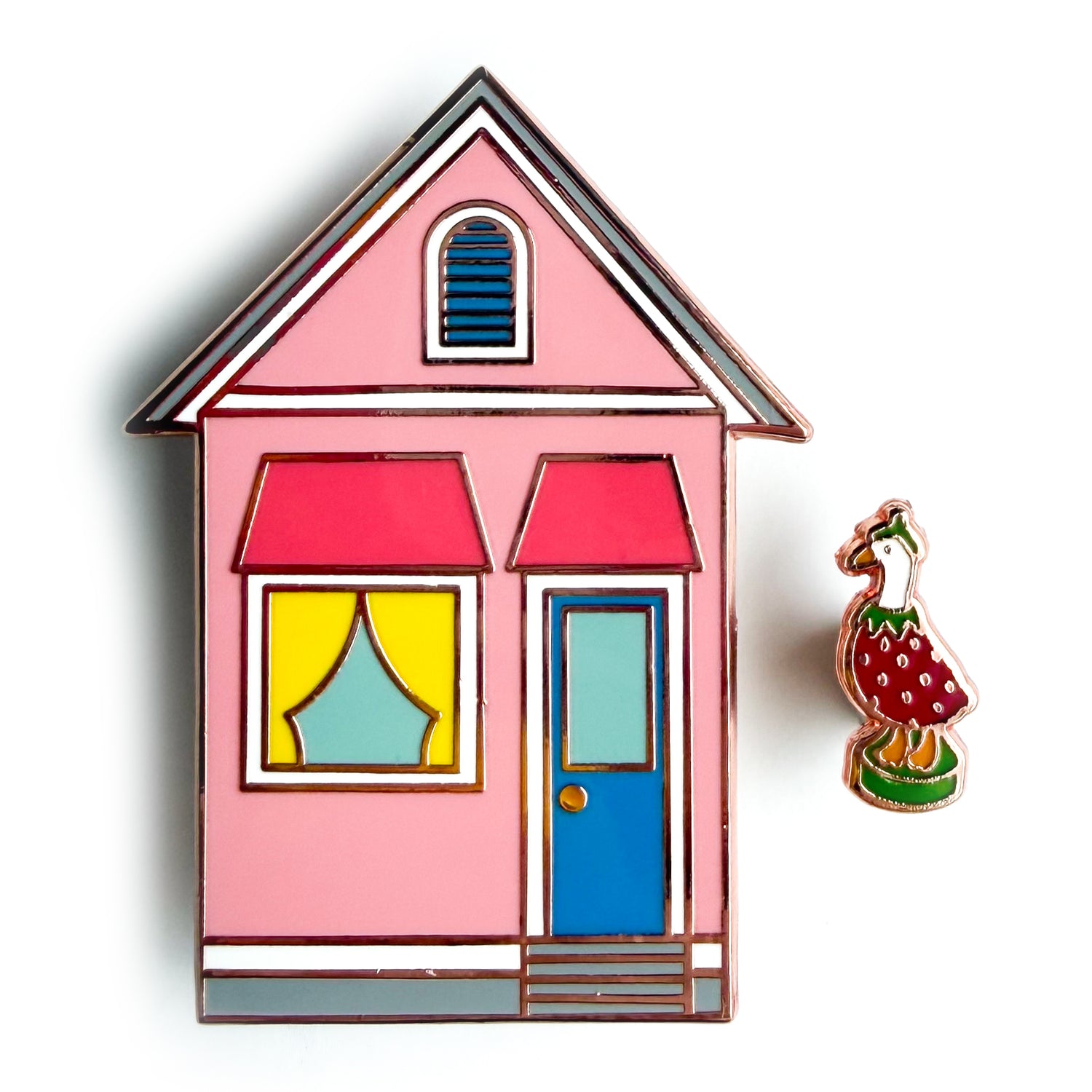 A pink house shaped enamel pin with a blue door and yellow curtains and a mini pin shaped like a porch goose in a berry costume. 