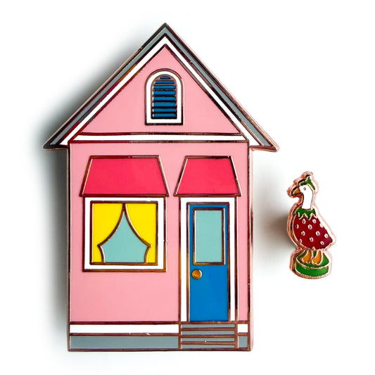 A pink house shaped enamel pin with a blue door and yellow curtains and a mini pin shaped like a porch goose in a berry costume.