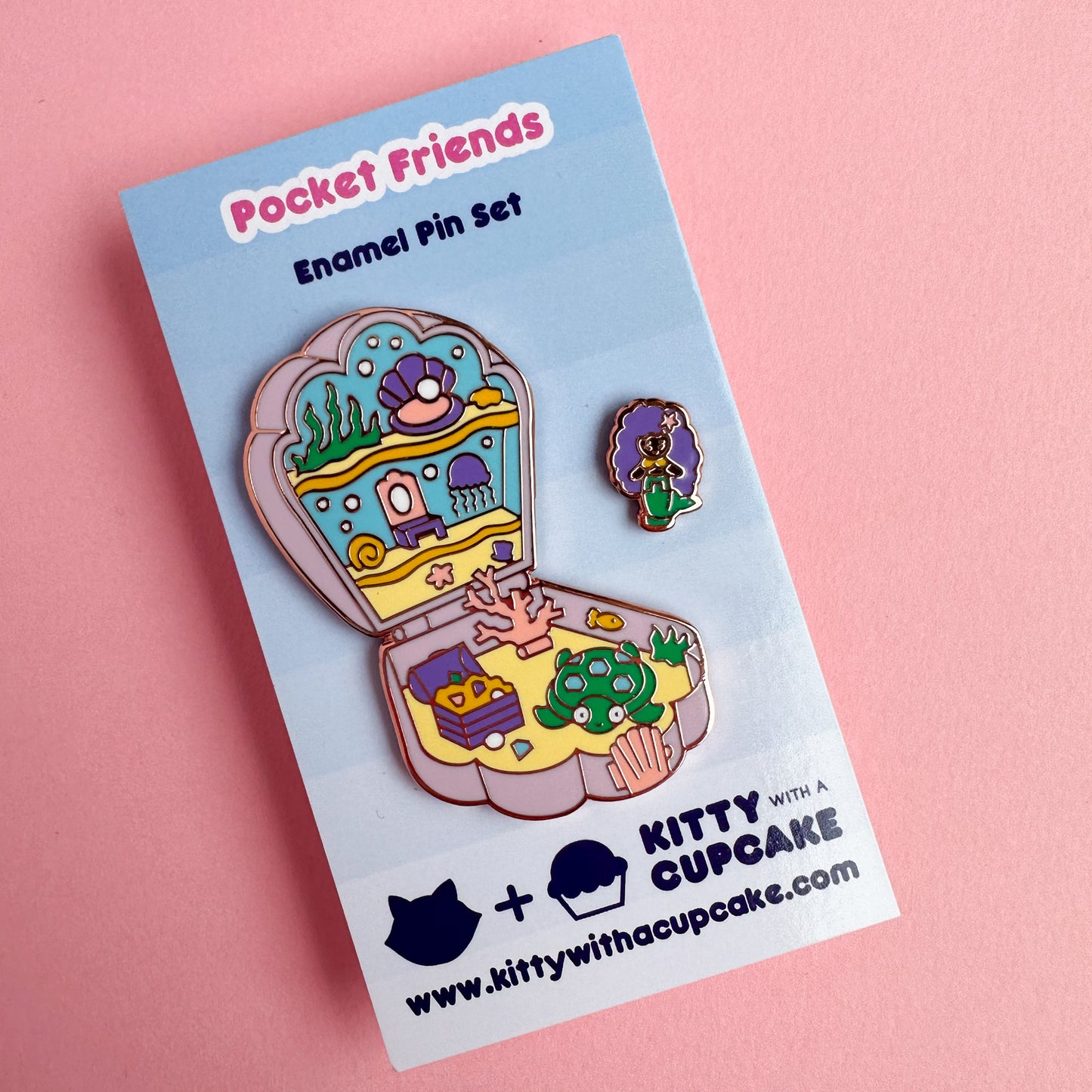 An enamel pin in the shape of a Polly Pocket compact shell with a mermaid mini pin. The shell compact is the mermaid's home with a jellyfish, coral, treasure chest, and a turtle. The pins are packaged on a blue card that reads "Pocket Friends Enamel Pin Set"