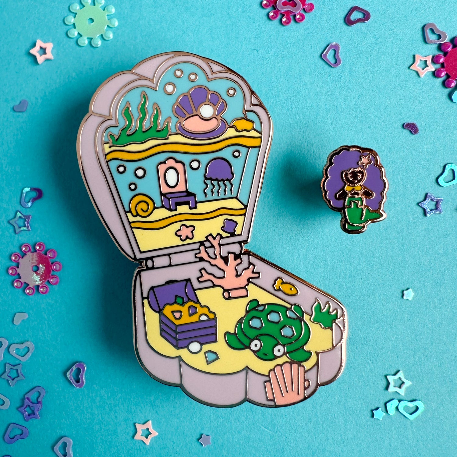 An enamel pin in the shape of a Polly Pocket compact shell with a mermaid mini pin. The shell compact is the mermaid's home with a jellyfish, coral, treasure chest, and a turtle. The pins are on a blue background with confetti