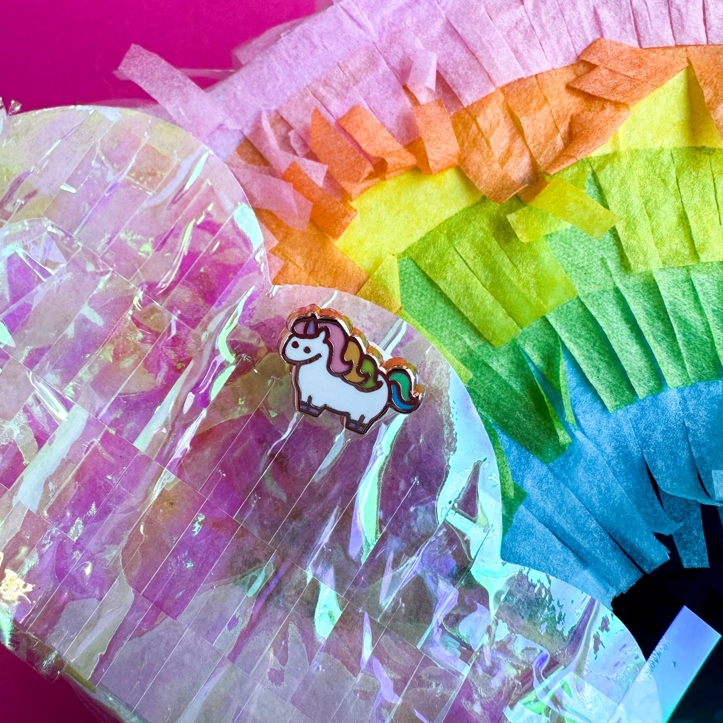 A mini enamel pin of a pastel rainbow unicorn on top of a pastel rainbow shaped piñata. 
