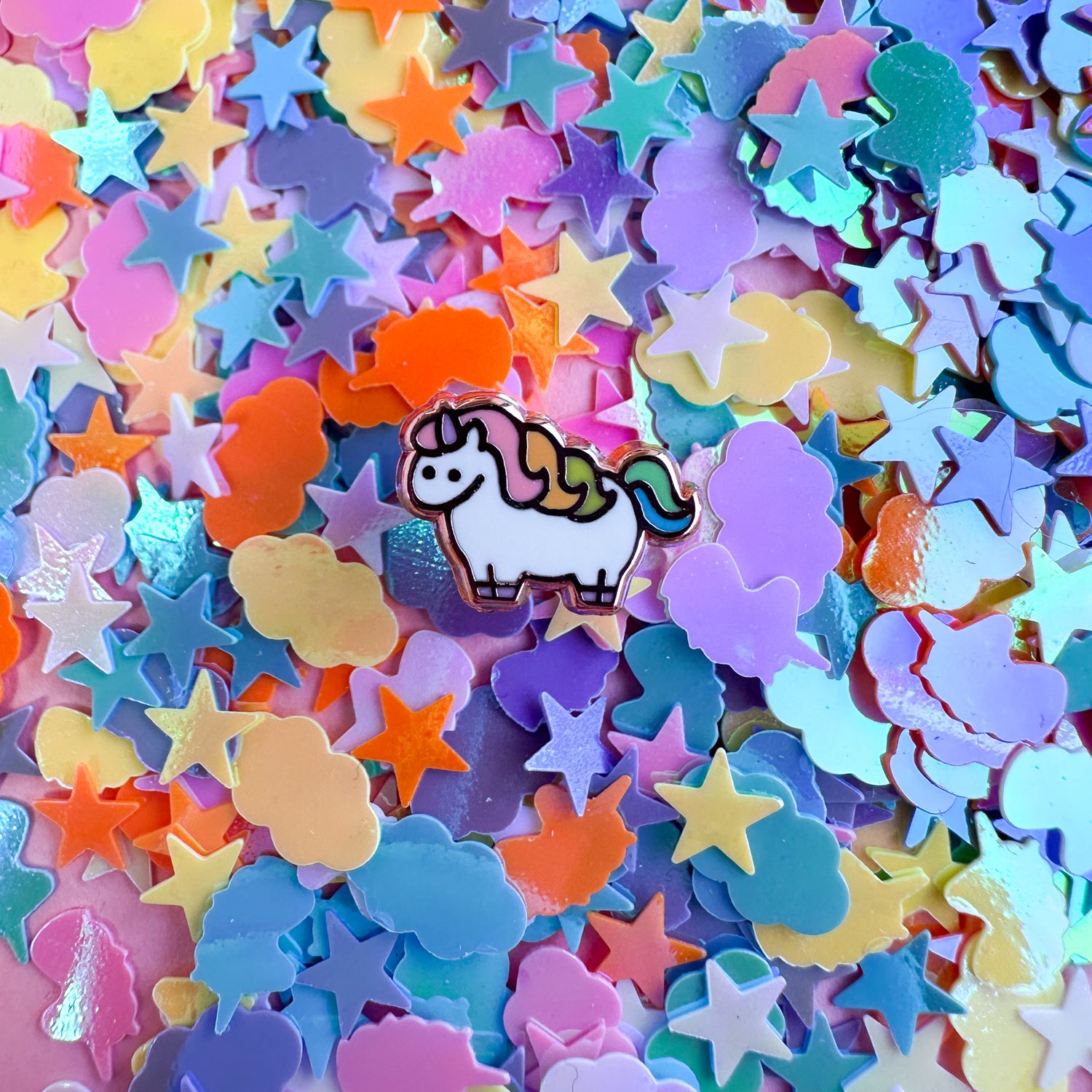 A cute mini unicorn enamel pin surrounded by pastel confetti shaped like clouds, stars, and unicorns. 