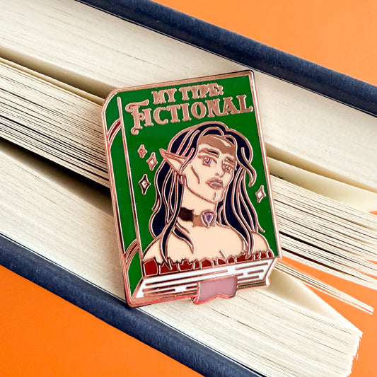 An enamel pin depicting a green book with the title “My Type: Fictional” written in gold. There is a hot androgynous elf on the cover with long black hair, big purple eyes, and luscious pink lips. They have sharp features, high cheek bones, and a chiseled jaw. They are wearing a gold crown, a gold choker with a purple gem, and an off-the-shoulder brown top. The book has gold accents on the spine, white sparkles on the cover, and a pink bookmark sticking out of the bottom. The pin is being on the splayed edg