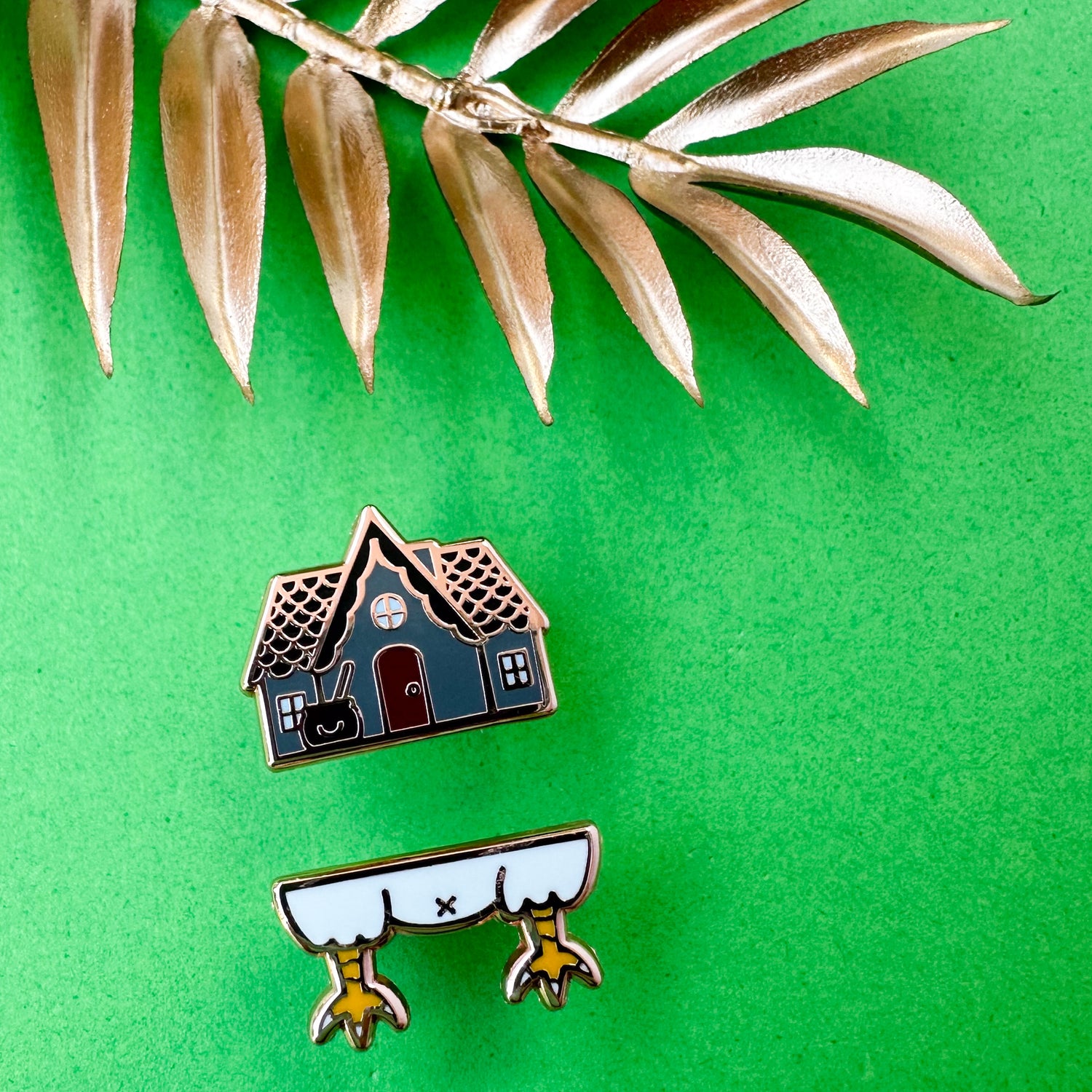Two baba Yaga's house pins, the top pin is a grey house with a cauldron and the bottom is white chicken legs. The pins are on a green background with gold leaves.
