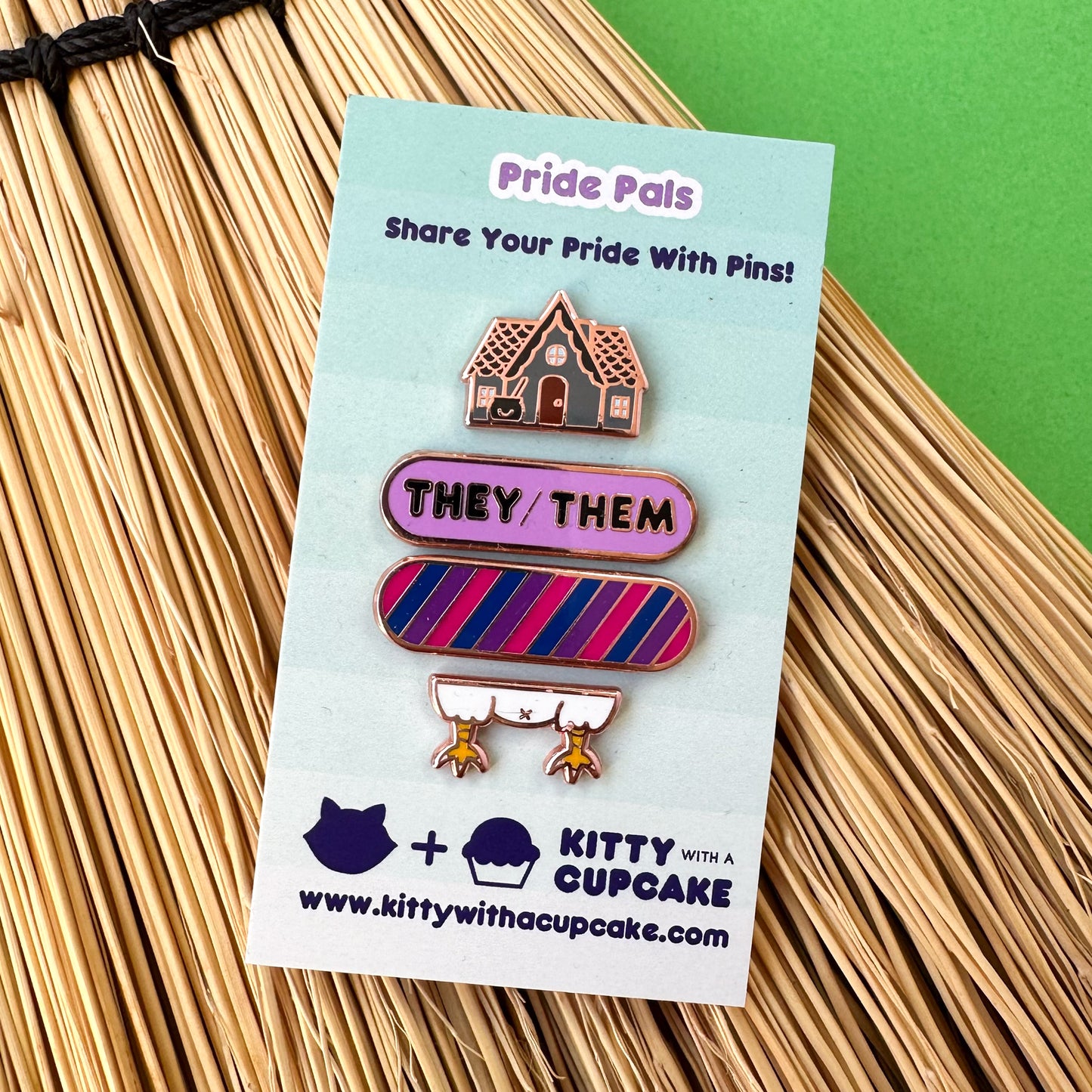 A card that reads "Pride Pals" with four pins on it in a stack, there is a grey house with a cauldron, a purple bandaid shaped pin with the words "They/Them" on it and a bandaid shaped pin in the colors of the bi pride flag, and the bottom pin is chicken legs. The card is on top of broom straw on a green background.