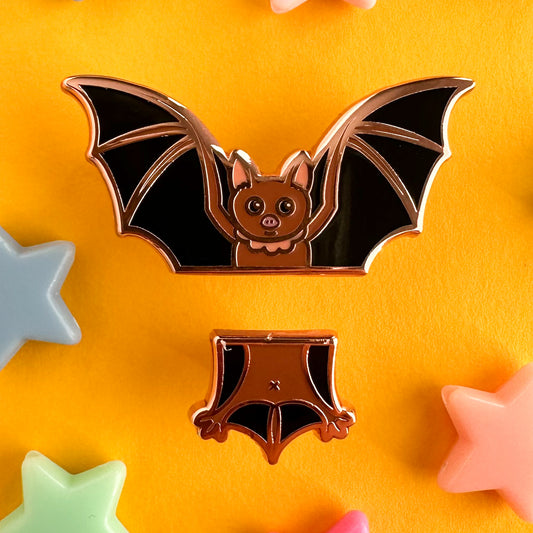 A set of enamel pins that come together to form a cute illustration of a bat, the top pin is the head and wings, the bottom is the tail and legs. The pins are on a yellow background with plastic pastel star beads around them.