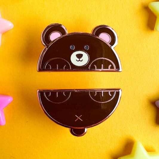 Two enamel pins that come together to form a bear cut in half horizontally. The top pin is the bear's head and front paws and the bottom is his tail and back paws.  The pins are on a yellow paper background.