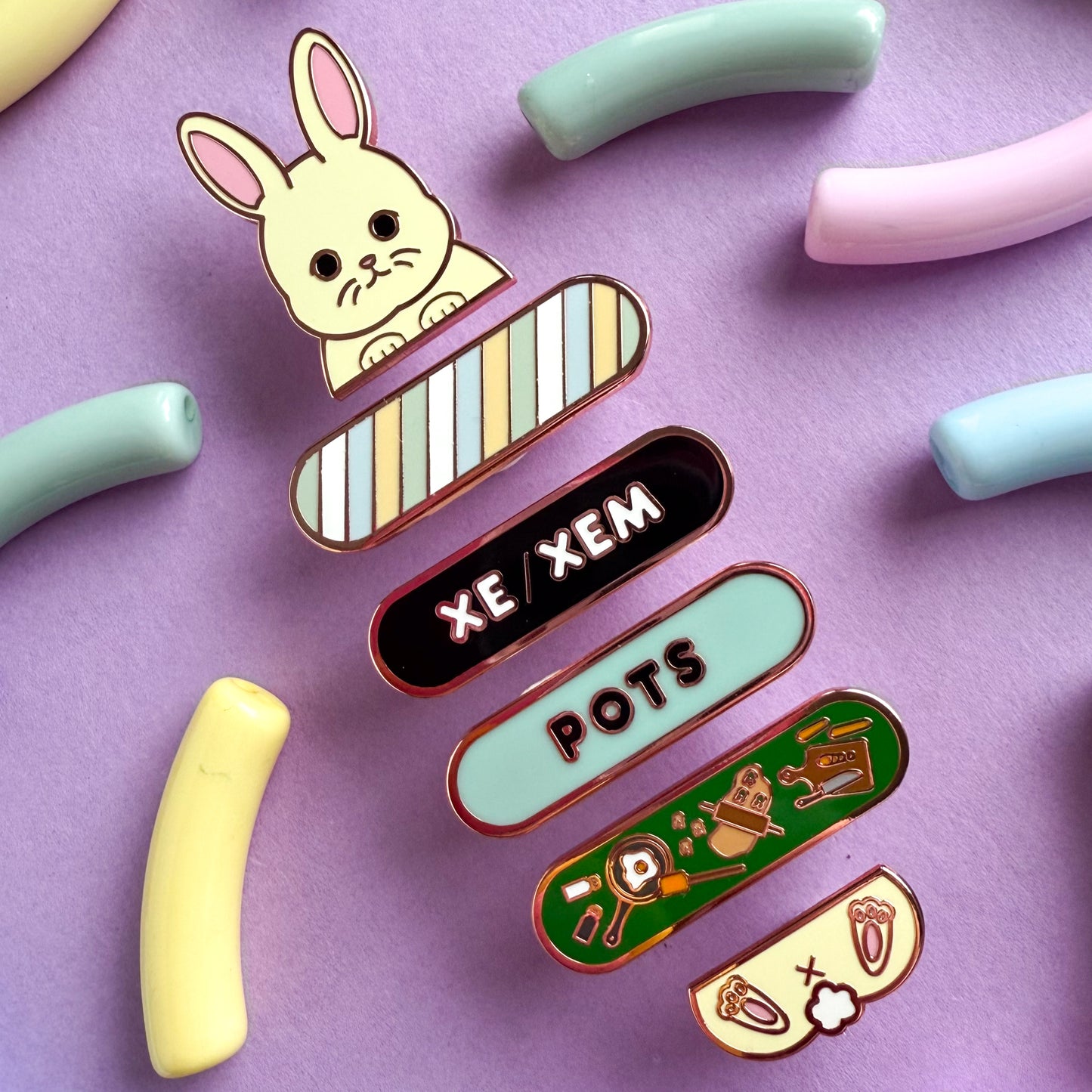 A stack of pride pals on a purple background, laid diagonal across the photo. This stack includes a bunny pal, an unlabeled plaque, a xe/xem plaque, a POTS plaque, and a cooking plaque.