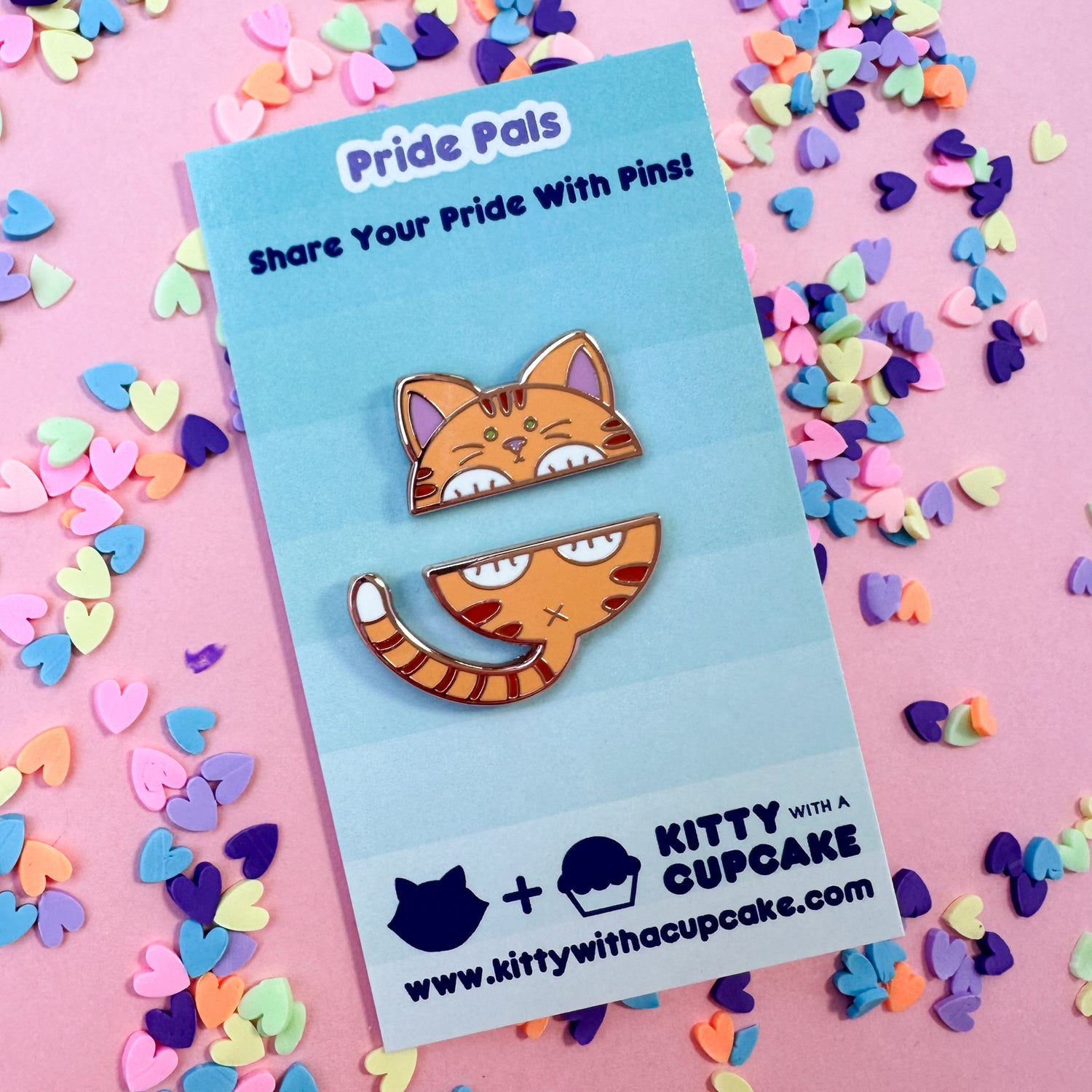 Two enamel pins that come together to form the top and bottom half of a cat packaged on a card. The background is pink with candy hearts around. 
