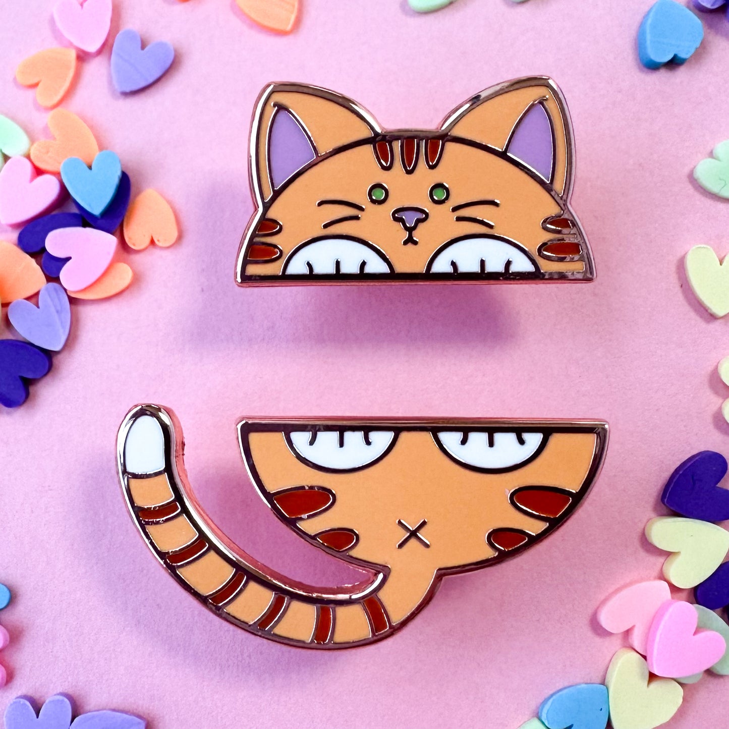 Two enamel pins that come together to form the top and bottom half of a cute cat. The cat is orange with stripes. The pin is on a pink background with candy hearts surrounding it. 