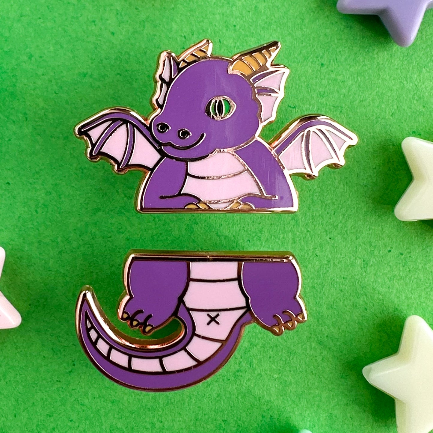 Two enamel pins that come together to form a cute cartoon dragon split in half horizontally. The top pin is the head, wings and arms. The bottom half is the legs and tail. The dragon is purple with a green eye and yellow horns. The pins are on a green backdrop with plastic star beads around them. 