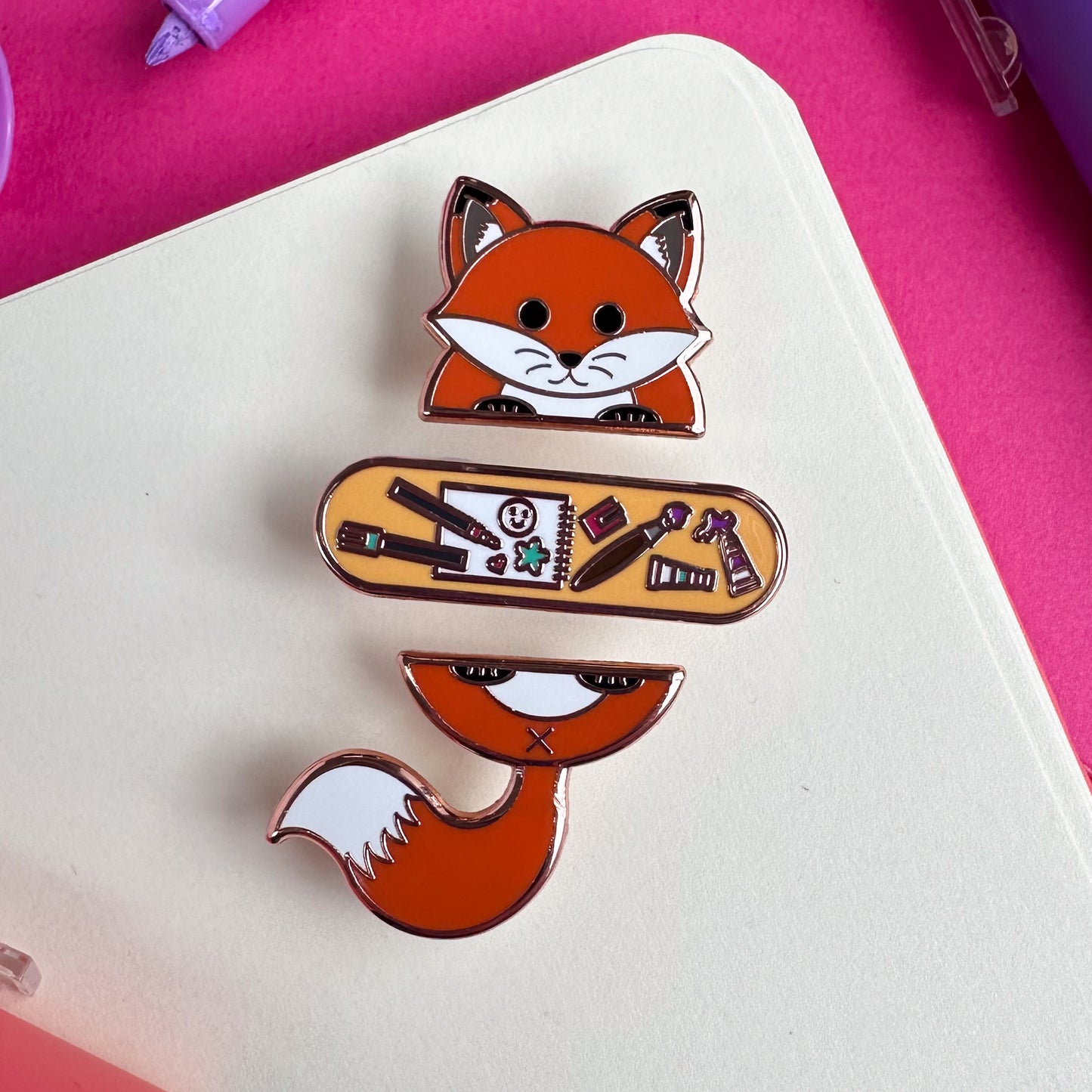 A set of enamel pins that form the top and bottom half of a fox holding a capsule shape that has illustrations of art supplies on it. The pins are on a sketchbook. 