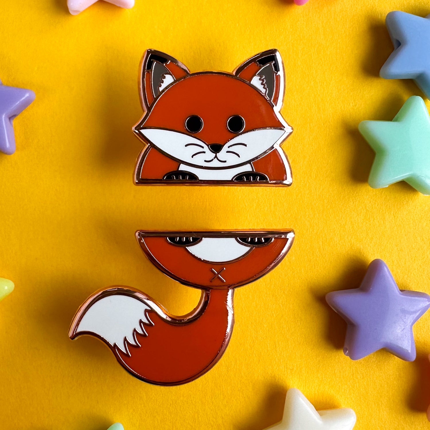 A set of enamel pins that comes together to form the top and bottom half of a fox. The top pin is the head and front feet of the fox and the bottom pin is the tail and the back feet of the fox. The pins are on a yellow background with star beads around them. 