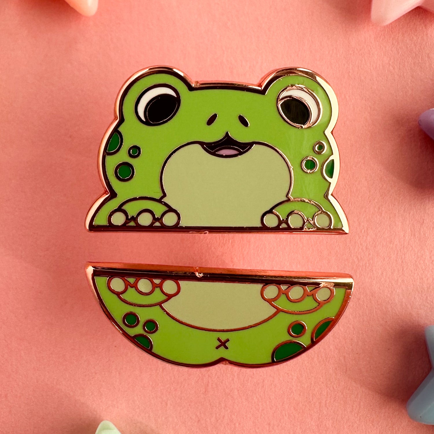 Two enamel pins that form the top and bottom half of a cute cartoony green frog with dark green spots. The pins are on a pink background.