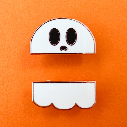 A set of two enamel pins that come together to form a ghost, one is the head and face and the other is the ghostly bottom. The pins are on an orange background. 
