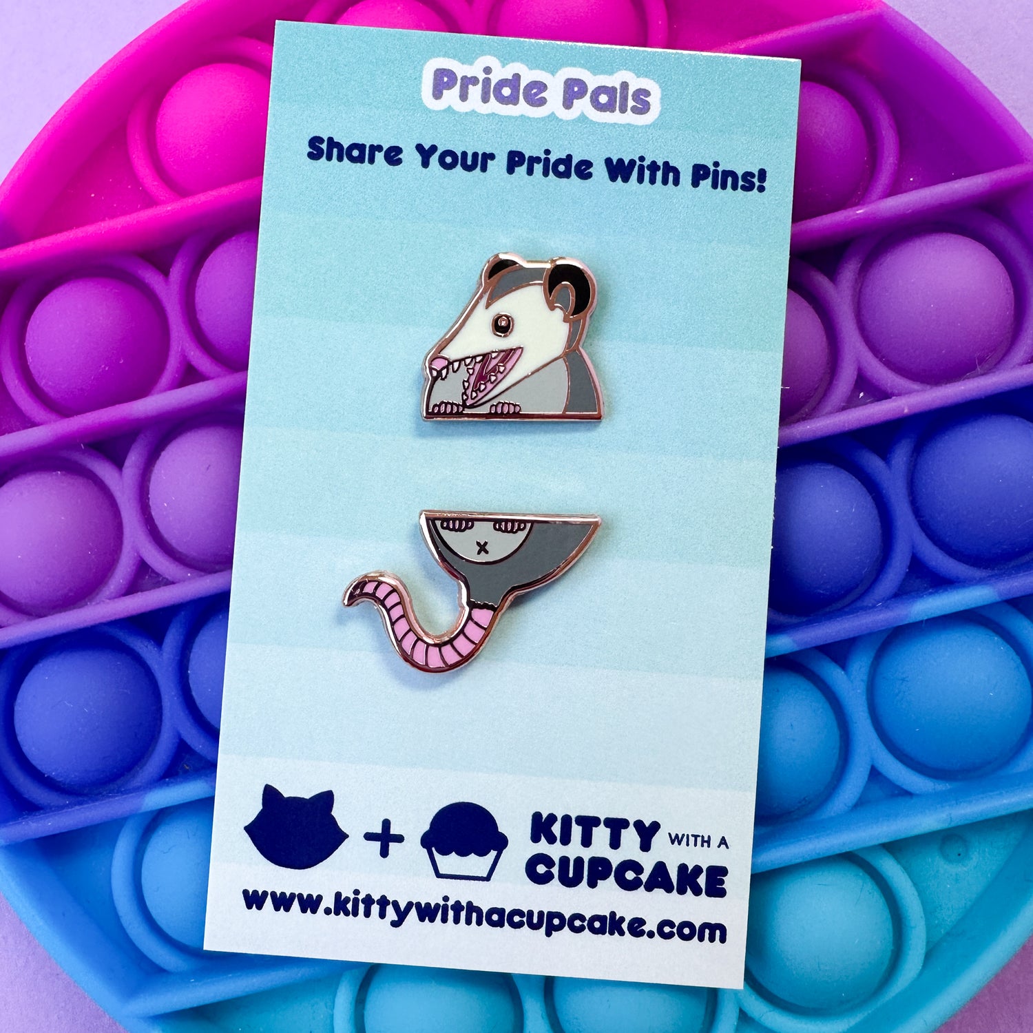 Two pins that form a possum that are packaged on a card that reads "Pride Pals" the card is on top of a fidget pop toy. 