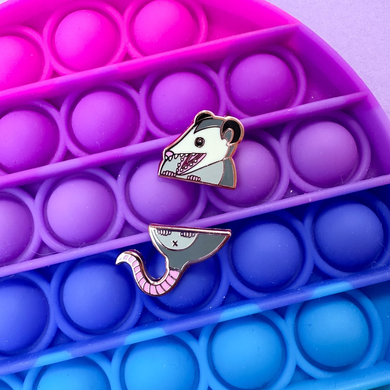 Two enamel pins that come together in the middle to form an opossum. The pins are resting on top of a fidget pop toy. 