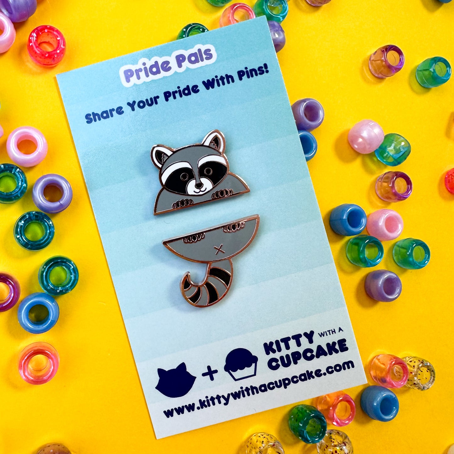 Two enamel pins that come together to form a cute raccoon. The pins are packaged on a card that is sitting on a yellow background with pony beads around it. 
