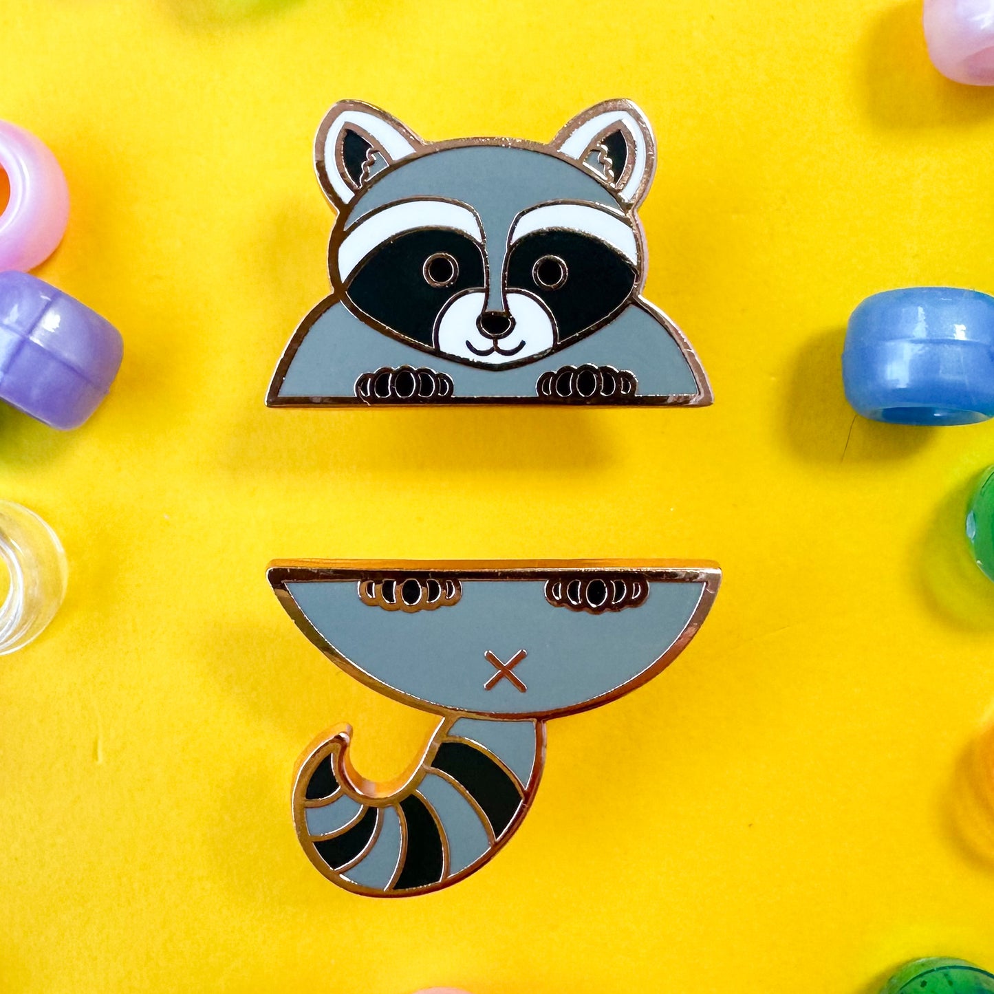 Two enamel pins that form a cute raccoon illustration, the pins are on a yellow background with pony beads scattered around it.