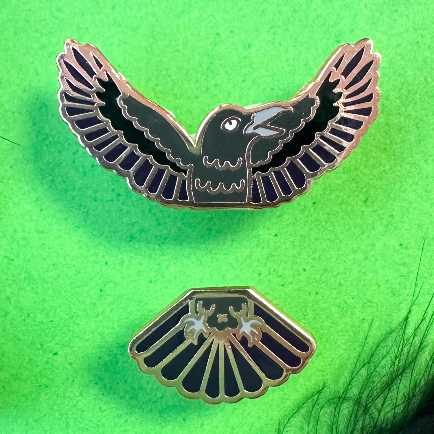 Two pins that come together in the middle to form a raven. The pin is on a green background with feathers in one corner. 