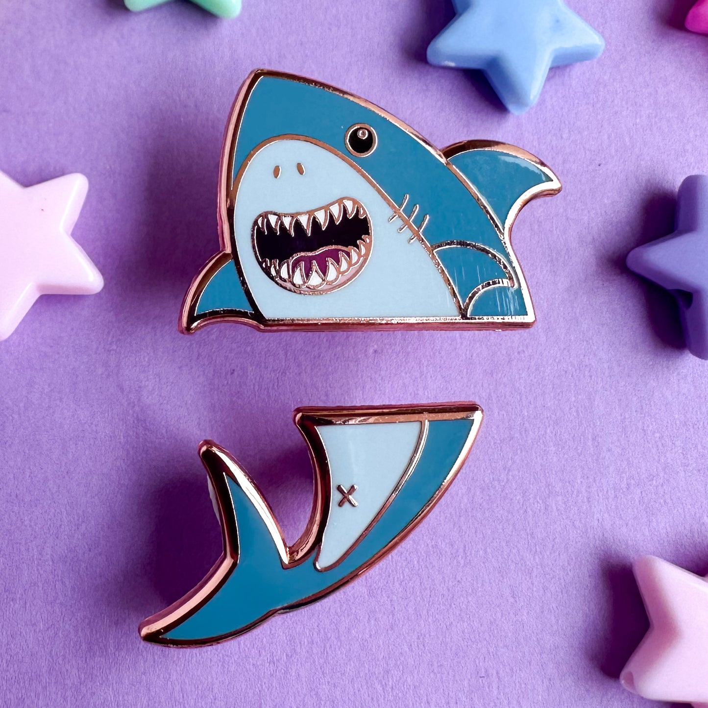 A pair of enamel pins that come together to form a cute illustration of a shark that is split horizontally across the middle. The top pin is the head and side fins, the bottom is the tail. The pins are on a purple paper background with plastic pony beads on it. 