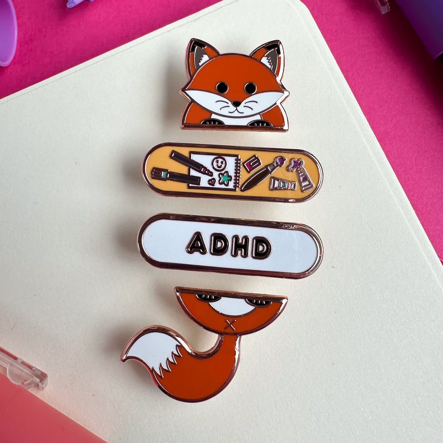 A set of enamel pins, the top pin is a cute cartoony fox head, underneath is a capsule shaped pin with art supplies on it, and below that is a white pin with the letters ADHD, the bottom pin is the tail and and feet of the fox. All these pins are on top of a sketchbook corner. 