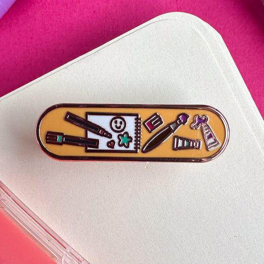 A capsule shaped enamel pin with cute illustrations of art supplies like paints, markers, and a sketchbook on it. The pin itself is on top of a sketchbook.
