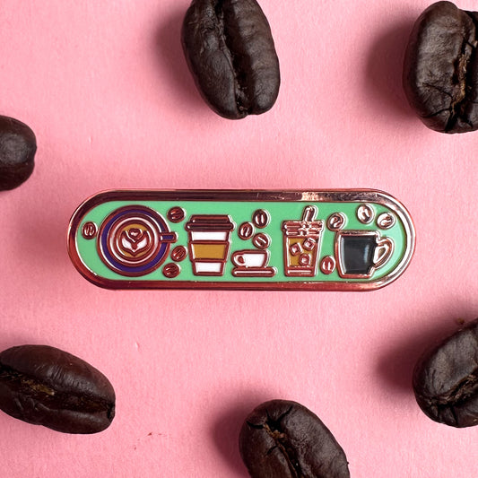 A capsule shaped enamel pin with illustrations of different coffee drinks on it including a latte with latte art, to-go hot drink, espresso cup, iced coffee and coffee mug. There are little coffee beans around these drinks. The pin is on a pink background with with real coffee beans around it.
