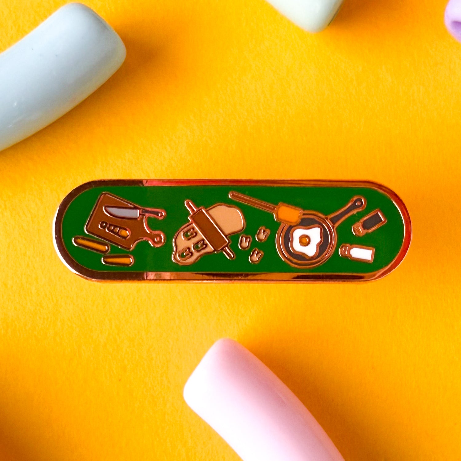 A bandaid shaped enamel pin with cooking themed illustrations. The pin is on a dark yellow background.