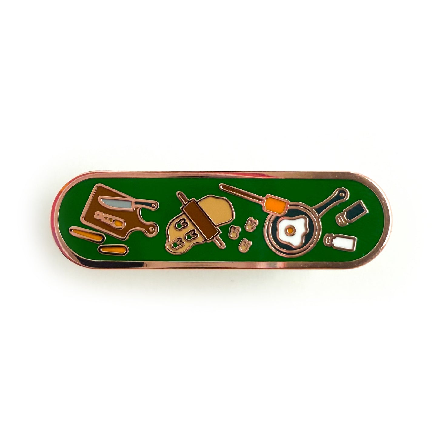 A bandaid shaped enamel pin with cooking themed illustrations on a deep green background. There is a cutting board with carrots, a rolling pin with cookie dough, a frying pan frying an egg, and salt and pepper shakers.