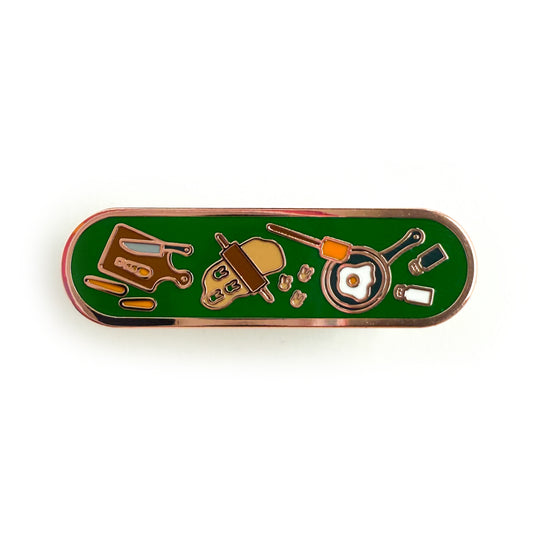 A bandaid shaped enamel pin with cooking themed illustrations on a deep green background. There is a cutting board with carrots, a rolling pin with cookie dough, a frying pan frying an egg, and salt and pepper shakers.