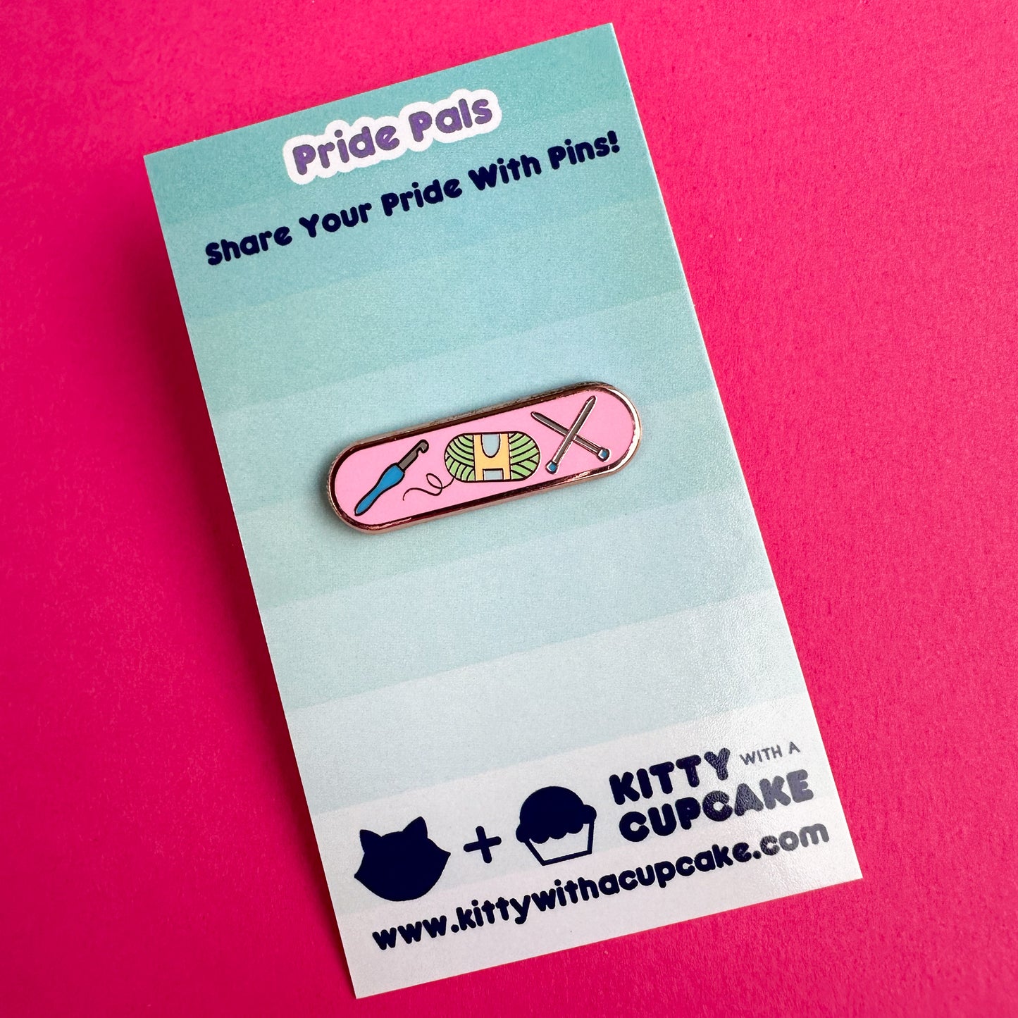 An enamel pin with illustrations of a crochet hook, yarn ball, and knitting needles on it. The pin is packaged on a card and the card is sitting on a hot pink background. 