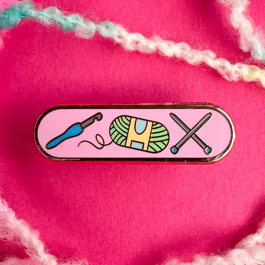 A bandaid shaped pin with a pink background and illustrations of a crochet hook, yarn ball, and knitting needles on it. There is boucle yarn around it and the pin is on a pink background. 