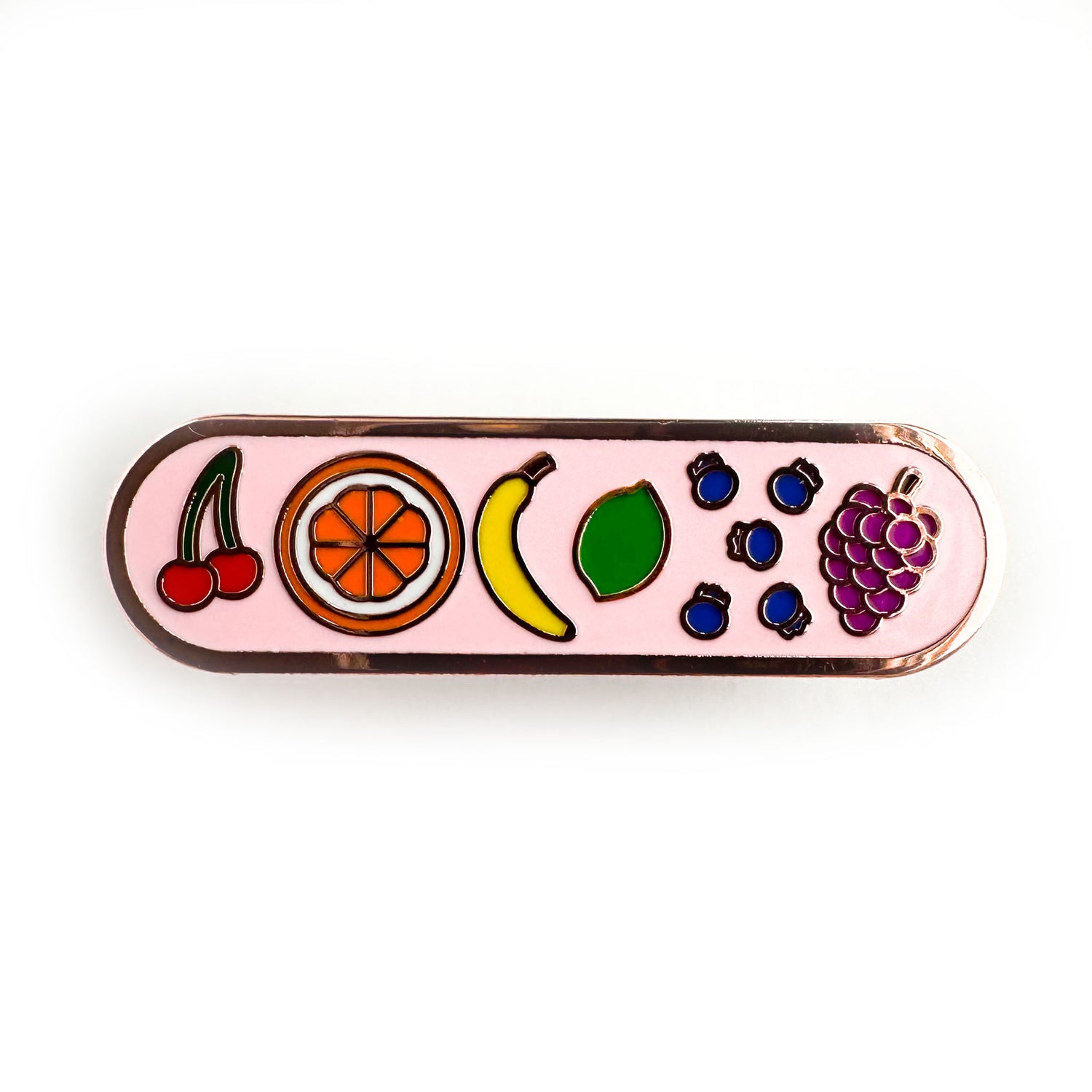 A capsule shaped enamel pin with little doodles of fruit on it arranged in rainbow order. There are cherries, an orange, a banana, a lime, blueberries, and grapes. The background of the pin is light pink. 