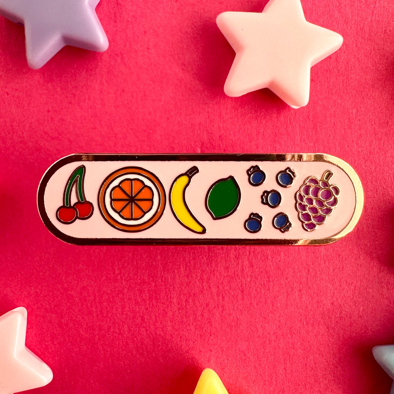 A capsule shaped enamel pin with little doodles of fruit on it arranged in rainbow order. There are cherries, an orange, a banana, a lime, blueberries, and grapes. The background of the pin is light pink. The pin is on a hot pink backdrop with plastic star beads around it. 