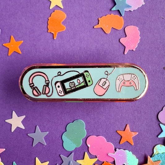 An oval shaped pin with illustrations of video game system equipment on it. The pin is on a purple background with pastel unicorn and star glitter around it. 