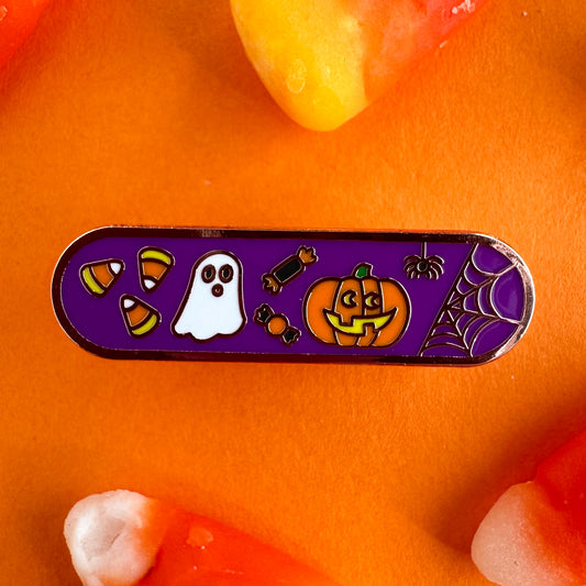 A capsule shaped enamel pin with a purple background and cute illustrations of candy corn, a ghost, candy, a jack o lantern, and a spider on it.  The pin is on an orange paper backdrop surrounded by candy corn.