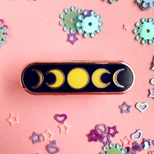 An oval pin with a full moon in the center and crescent moons framing it. The pin is on a pink background with glitter around it. 