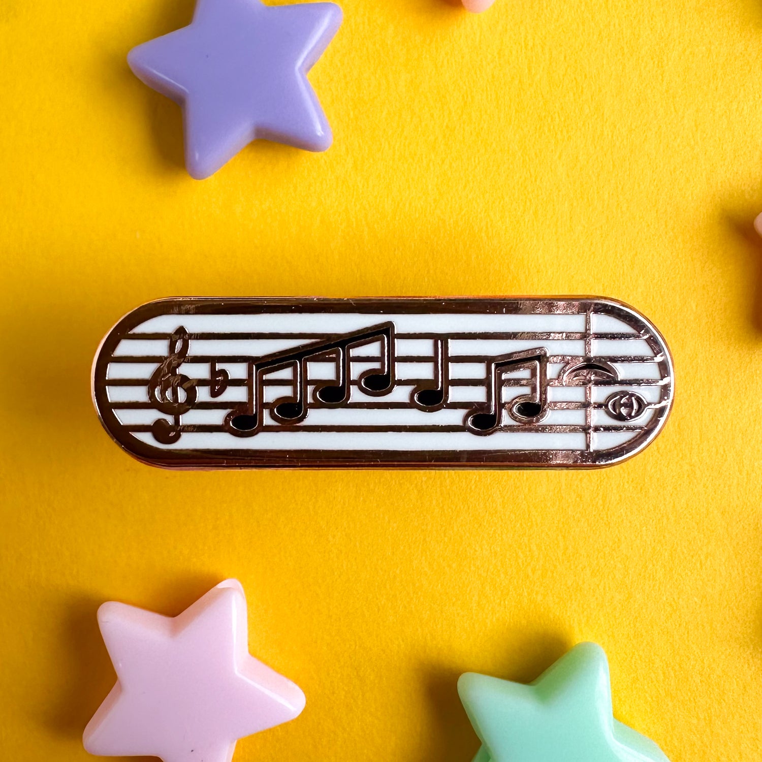 A music staff on a capsule shaped enamel pin. The pin is on a yellow background with pastel star beads around it. 