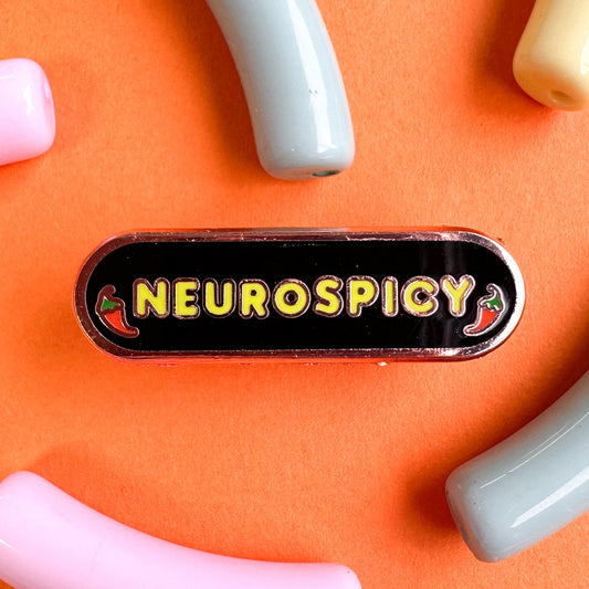 An enamel pin with the word "Neurospicy" and chili peppers around it. The pin is on an orange background with pastel beads around it. 